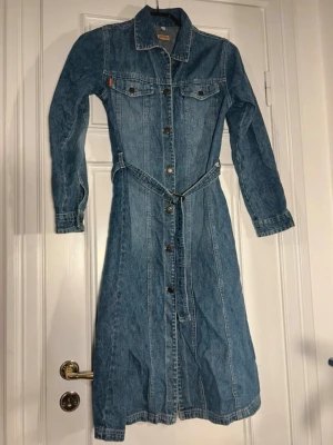 Lång jeansklänning med bälte Levi's - Snygg lång jeansklänning från Levi's i klassisk blå denim. Klänningen har långa ärmar, knappar hela vägen framtill, två bröstfickor med lock och ett matchande bälte i midjan. Perfekt för dig som gillar retro och tidlös stil.