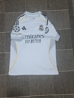 Real Madrid matchtröja Adidas vit - Säljer en vit Real Madrid matchtröja från Adidas med grå och gula detaljer. Tröjan har klubbmärke, Adidas-logga och sponsortryck på bröstet samt Champions League-märke och UEFA Foundation-märke på ärmen. Tillverkad i lätt och ventilerande material.