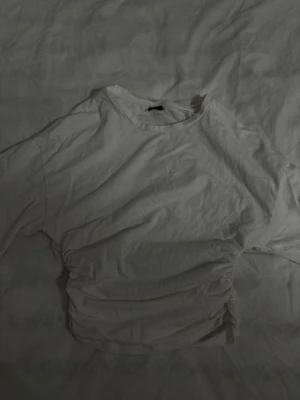 Vit kortärmad t-shirt från gina tricot - Säljer en vit t-shirt från gina tricot med korta ärmar och rund hals. T-shirten har en enkel och clean look med lätt rynkade detaljer på sidorna som ger en snygg twist. Tillverkad i mjuk bomull som känns skön mot huden. Små i storleken passar folk med xs 