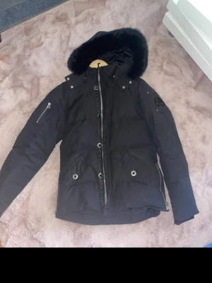 Svart dunjacka med päls från Moose Knuckles - Säljer en svart dunjacka från Moose Knuckles med avtagbar huva och svart päls. Jackan har två stora fickor med knappar framtill, en ärmficka med dragkedja och logotyp på ena ärmen. Perfekt för kalla vinterdagar.