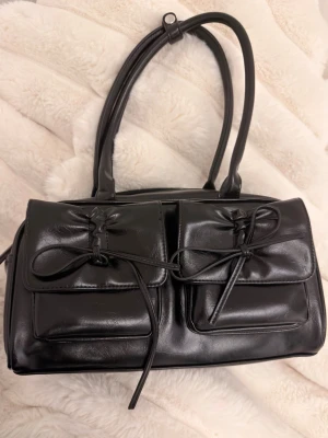 Svart axelväska med dubbla fickor even & odd - Snygg svart axelväska i konstläder med två stora framfickor som stängs med dekorativa knytband. Väskan har dubbla handtag och ett rymligt huvudfack med dragkedja. Perfekt för dig som gillar stilrena detaljer och praktiska fack. Använd 1 gång köpt på Zalando.