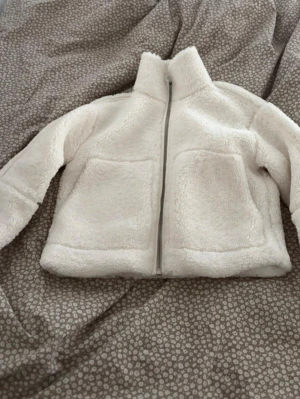 Vit teddyjacka med dragkedja - Mysig vit teddyjacka med hög krage och hel dragkedja framtill. Jackan har två stora framfickor och är gjord i fluffigt, mjukt material som ger en riktigt chill vibe. Perfekt för kalla dagar när du vill vara både varm och stylish.