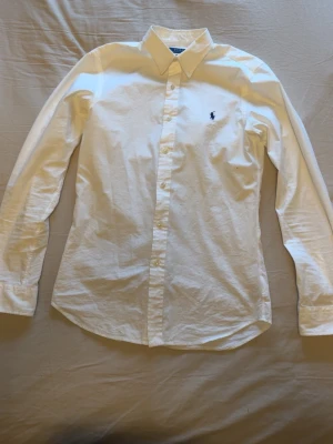 Vit skjorta från Polo Ralph Lauren - Klassisk vit skjorta från Polo Ralph Lauren med broderad logga på bröstet. Skjortan har lång ärm, knappar framtill och en snygg krage. Perfekt till många olika stilar och enkel att matcha.