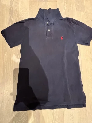 Mörkblå piké från Polo Ralph Lauren - Klassisk mörkblå pikétröja från Polo Ralph Lauren med röd broderad logga på bröstet. Tröjan har korta ärmar, krage och knappar vid halsen. Tillverkad i mjuk bomull och har en tidlös passform som funkar till allt.