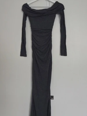 Mörkgrå offshoulder långklänning från Bershka - Snygg mörkgrå långklänning med offshoulder-design och långa ärmar från Bershka. Klänningen har rynkade detaljer längs sidorna och en slits framtill för extra stilpoäng. Tillverkad i mjukt och stretchigt material som sitter bekvämt och följer kroppen.