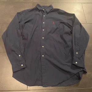 Mörkblå skjorta från polo ralph lauren sport golf - Klassisk mörkblå skjorta från polo sport med röd broderad logga på bröstet. Skjortan har lång ärm, knappar framtill och button down-krage. Tillverkad i mjuk bomull och passar perfekt till en stilren look. Passformen är L/XL