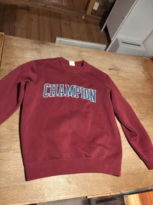 Vinröd sweatshirt från Champion - Snygg vinröd sweatshirt från Champion med stort tryck över bröstet i vitt och blått. Klassisk rund halsringning och långa ärmar. Tillverkad i mjuk bomullsmix som är skön att ha på sig. Funkar för er som är ligger mellan S och M. Fråga gärna om fler bilder eller om ni har några frågor