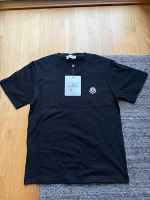 Svart Moncler t-shirt - 399kr! Köp båda för 649! Snygg svart t-shirt från Moncler med klassisk rund hals och korta ärmar. Tillverkad i mjuk bomull och pryds av Monclers ikoniska logotyp på bröstet. Perfekt basplagg för en clean och stilren look.
