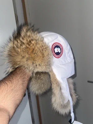 Vit Canada Goose trappermössa - Säljer en vit trappermössa från Canada Goose med stora öronlappar och äkta pälsdetaljer. Mössan har det klassiska Canada Goose-märket på sidan och knytband under hakan. Perfekt för kalla vinterdagar och riktigt snygg till vinterjackan. Köpt i Amsterdam på canada goose kvitto kan letas upp inte 100% att jag hittar! L/XL jag som har S passar den och är ytterst lite stor men sitter en då! Väldigt omhändertagen inget anmärkningsvärt päls i topp klass!