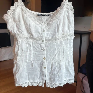 Vit spetsdetaljerad linnetopp från Zara - Superfin vit linnetopp från Zara med spetsdetaljer längs axelband, halsringning och nederkant. Toppen har knappar framtill och tunna axelband. Materialet är linne och toppen har en romantisk vibe med broderade detaljer och v-ringad hals.