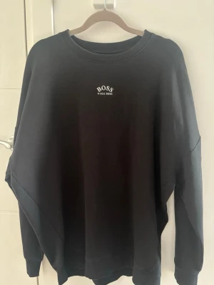 Svart sweatshirt från Hugo Boss - Svart sweatshirt från Hugo Boss med liten vit logga framtill och stor, stilren BOSS- och HUGO BOSS-tryck på ryggen. Tröjan har rund halsringning. Perfekt för en clean och trendig look.