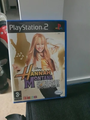Hannah Montana Spotlight World Tour PS2 - PlayStation 2-spelet Hannah Montana Spotlight World Tour med blå plastfodral. Omslaget har färgglad design med musiktema och bilder på Hannah Montana. Spelet är för 3+ år och har stöd för flera språk. Perfekt för dig som gillar musikspel och Disney.