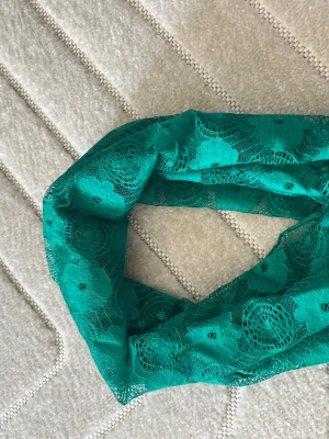 Grön spetsig tubsjal - Snygg grön tubsjal i spets med blommigt mönster. Sjalen är gjord i ett tunt, genomskinligt material som ger en elegant känsla och passar perfekt för att lyfta din outfit. Enkel att styla och ger en cool touch till din look.