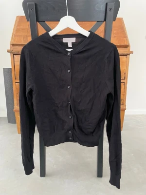 Svart kofta från H&M, storlek S - Klassisk svart kofta från H&M med rund halsringning och knäppning framtill. Långärmad modell i mjukt, tunt material som passar perfekt till lager på lager. Enkel och stilren design som funkar till det mesta.