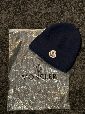 Marinblå Moncler mössa. - Säljer en marinblå ribbstickad mössa från Moncler med klassisk logga framtill. Mössan är i mjuk ull och har vikt kant för extra stilpoäng. Perfekt accessoar för kyliga dagar och snygg till alla outfits. Jättebra skick. Pris kan disktuteras. Hör av er vid frågor eller funderingar.