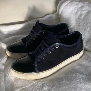 Mörkblå Lanvin sneakers med lackdetaljer - Snygga mörkblå sneakers från Lanvin med ovandel i mocka och tåparti i blankt lack. Skorna har vita gummisulor och matchande mörkblå skosnören. Klassisk låg modell med stilrena detaljer och metallöljetter. Storlek 6 som motsvarar 41-42. Bara skriva om ni har frågor!☺️