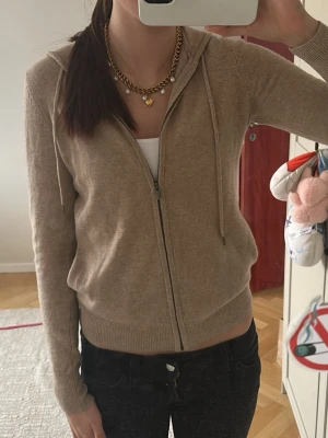 Beige stickad hoodie med dragkedja - Mysig beige stickad hoodie med dragkedja framtill och huva med dragsko. Tröjan har långa ärmar, ribbade muddar och nederkant samt två fickor fram. Perfekt för lager på lager och enkel att matcha med olika outfits.