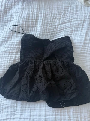 Svart broderad topp från Zara - Supersöt svart topp från Zara med virkad överdel och broderad, volangig nederdel. Toppen har en axelbandslös design med twistad detalj framtill och fina hålbroderier på tyget. Perfekt för dig som vill ha en unik och trendig look.