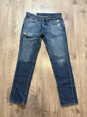 Hollister - Säljer ett par blå jeans från Hollister med raka ben och slitningar på låret och vid fickan. Jeansen har klassisk femficksmodell, knappgylf och är gjorda i bomull med en snygg tvättad look. Perfekta för dig som gillar en avslappnad och trendig stil.