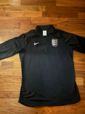 Svart England Nike träningströja - Snygg svart långärmad träningströja från Nike med Englands landslagsmärke på bröstet och halv dragkedja framtill. Perfekt för fotbollsträning eller som sportig vardagsstil. Tillverkad i ett lätt och funktionellt material.