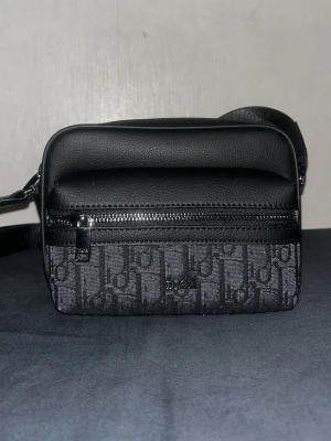 Svart axelväska från Dior - Snygg svart axelväska från Dior med klassiskt monogrammönster på framsidan och detaljer i svart läder. Väskan har en justerbar axelrem och en praktisk dragkedja framtill. Perfekt för dig som vill ha en stilren och trendig accessoar.