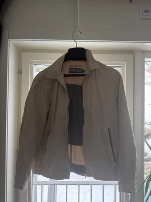 Beige vindjacka från Joy - Stilren beige vindjacka från Joy med klassisk krage och dragkedja framtill. Jackan har två snedställda fickor med dragkedja och elastisk nederkant. Insidan har meshfoder för extra komfort. Perfekt för vår och höst.