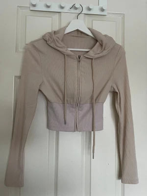 Beige cropped hoodie med dragkedja - Trendig beige cropped hoodie med dragkedja framtill och långa ärmar. Tillverkad i ribbat tyg och har en huva med dragsko. Perfekt för en avslappnad och cool stil.
