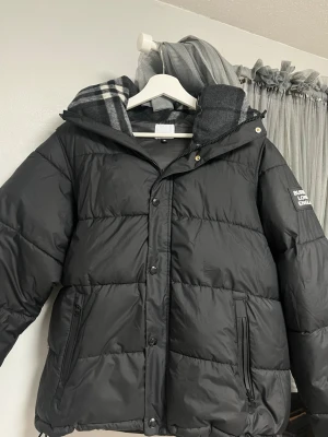 Burberry puffer I Väst - Säljer en svart pufferjacka från Burberry London England med huva och dragkedja framtill. Jackan har två stora fickor med dragkedja och en patch med logga på ärmen. Perfekt oversized look och riktigt skön för kalla vinterdagar.