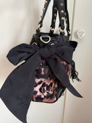 Juicy Couture väska - Väldigt gammal juicy couture väska som legat i en byrå utan användning, köptes begagnad för flera år sedan. Den har små skavanker men fortfarande fin, rosetterna kan knytas hur man vill. Fickor finns både inuti och utanpå väskan. Långt axelband medföljer till väskan.