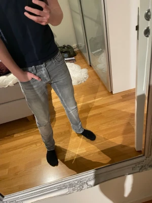 Grå slim fit jeans Jack N Jones - Säljer ett par grå slim fit jeans med klassisk femficksdesign och dragkedjegylf. Jeansen har en snygg tvättad look och sitter tajt längs benen. Perfekta för dig som gillar en modern och stilren stil. Modell på bilden är 172