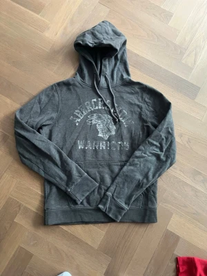 Abercrombie & Fitch hoodie - Snygg mörkgrå hoodie med tryck framtill från Abercrombie & Fitch. Ruggigt coolt tryck som ger hela plagget en archive känsla. Skick 7/10. Strl M. 