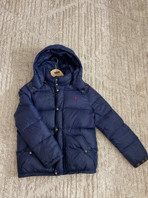 Mörkblå dunjacka från Polo Ralph Lauren - Snygg mörkblå dunjacka från Polo Ralph Lauren med avtagbar huva och guldiga knappar. Jackan har två snedställda fickor framtill och den klassiska röda loggan broderad på bröstet. Perfekt för kalla vinterdagar och riktigt skön att bära.