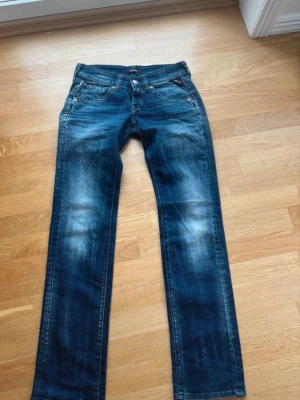 Blå raka jeans - Säljer ett par blå jeans med raka ben och snygga slitningar framtill och baktill. Jeansen har klassisk femficksdesign, knappgylf och kontrastsömmar. Materialet är denim i bomull. Perfekta för en avslappnad och trendig look.