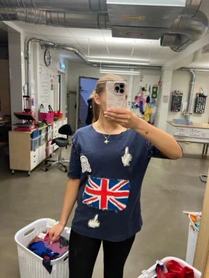 Mörkblå t-shirt med Union Jack och tryck - Unik mörkblå t-shirt med stort Union Jack-tryck framtill, coola patches och texten 'ROCK N FUCKING ROLL' på ryggen. T-shirten har korta ärmar och en avslappnad passform. Perfekt för dig som gillar en edgy och personlig stil.