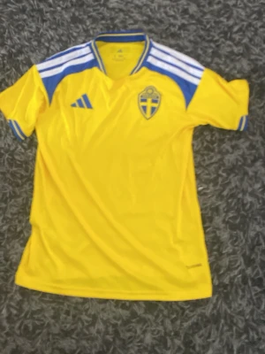 Svensk landslagströja Adidas gul/blå - Gul svensk landslagströja från Adidas med blå och vita detaljer, korta ärmar och broderat landslagsemblem på bröstet. Tillverkad i lätt och ventilerande material, perfekt för fotboll eller träning. Klassisk design med Adidas-logga och ränder på axlarna. ENDAST TESTAD