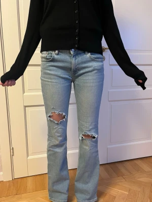 Ljusblå bootcut jeans - Säljer ett par ljusblå bootcut jeans med slitningar över båda knäna. Jeansen är midrise och är tillverkade i denim. Perfekta för en avslappnad och trendig look.