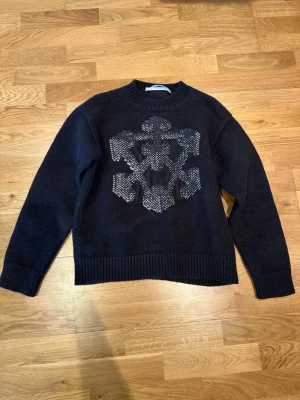 Svart stickad tröja från Off-White - Säljer en svart stickad tröja från Off-White med ett stort grått grafiskt mönster på bröstet och texten 'OFF' i grått på ryggen. Tröjan har rund hals, ribbade muddar och är gjord i ett tjockare material som känns riktigt lyxigt. Tag finns, samt screenahot på att den är äkta. Storlek L men skulle säga att den sitter mer som en M-S. 