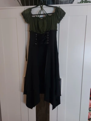 Grön och svart medeltidsklänning cosplay - Säljer en cosplayklänning i medeltidsstil med grön off-shoulder överdel och svart kjol med snörning framtill. Kjolen har asymmetrisk skärning och överdelen har puffärmar och axelband som korsas i ryggen. Perfekt för lajv, maskerad eller temafest.