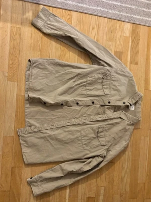 Beige skjorta med bröstfickor - Stilren beige skjorta med två bröstfickor och knappar framtill. Skjortan har lång ärm och klassisk krage, perfekt för en avslappnad men snygg look. 