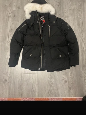 Svart dunjacka med pälskant moose  knukles - Snygg svart dunjacka från moose med stor huva och vit pälskant. Jackan har flera dragkedjeförsedda fickor, tryckknappar och en patch på ärmen. Insidan har ett coolt Moncler-märke. Perfekt för kalla vinterdagar.