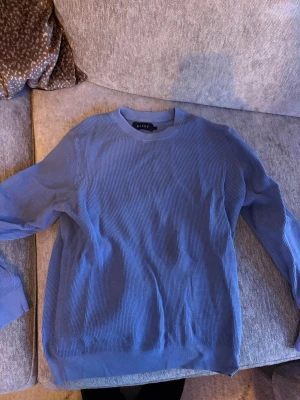 Blå stickad tröja från Riley - Säljer en stilren blå stickad tröja från Riley. Tröjan har rund halsringning, lång ärm och är tillverkad i ett tunt, mjukt material med diskret struktur. Perfekt att matcha med jeans eller kjol för en clean look.