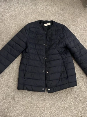 Svart pufferjacka med tryckknappar - Säljer en marinblå pufferjacka med raka sömmar och tryckknappar framtill. Jackan har en enkel, stilren design utan luva och är lätt vadderad för extra värme. Perfekt för kalla dagar och enkel att matcha med olika outfits.