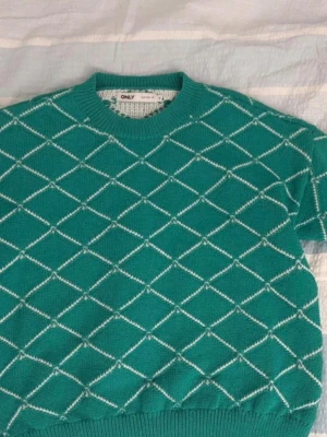 Grön stickad tröja från ONLY - Snygg grön stickad tröja från ONLY med vitt rutmönster. Tröjan har rund halsringning och ribbade muddar vid ärmslut och nederkant. Perfekt för dig som vill ha en trendig och bekväm look.