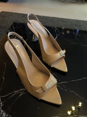 Helt oanvända klackar strl.38-40 - Snygga beige pumps i lackat material med spetsig tå och öppen häl. Skorna har en elegant slingback-rem . Klacken är smal och något utsvängd för en trendig look. Perfekta för att lyfta vilken outfit som helst. Klackarna är otroligt bekväma att gå i och inte för höga. Då klackarna är öppna vid hälen och tån så passar den strl 38-40. Nypris ca 800kr. Priset kan diskuteras.