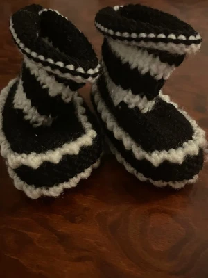 Svartvita virkade tofflor - Unika handgjorda tofflor i svart och vitt med virkade vågmönster. De har en högre skaftdesign och är gjorda i mjukt garn som känns skönt mot huden. Perfekta för att hålla fötterna varma hemma.