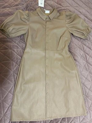 Beige skjortklänning från Vila - Trendig beige skjortklänning från Vila i syntetmaterial med puffärmar och knappar hela vägen framtill. Klänningen har klassisk krage och korta ärmar med resår vid ärmslut. Perfekt för dig som gillar stilrena och moderna plagg.