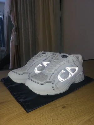 Vita Dior B30 sneakers med reflexdetaljer - Säljer ett par vita Dior B30 sneakers med feta reflexdetaljer på sidorna och meshpaneler för extra andningsförmåga. Skorna har chunky sula och klassisk snörning. Perfekta för dig som vill sticka ut med exklusiva och trendiga sneakers.