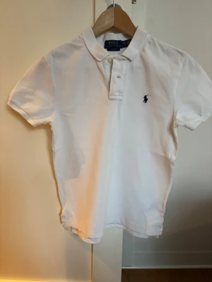 Vit pikétröja från Ralph Lauren - Klassisk vit pikétröja från Polo Ralph Lauren i slim fit-modell. Tröjan har korta ärmar, krage med två knappar och den ikoniska mörkblå logotypen broderad på bröstet. Tillverkad i mjukt bomullsmaterial som andas.