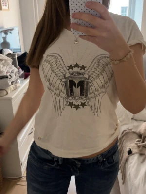 Vit t-shirt med vingar och stjärnor - Vit t-shirt med stort tryck av vingar, stjärnor och bokstaven M framtill. T-shirten har rund hals och korta ärmar. Perfekt för dig som gillar grafiska tryck och en avslappnad stil.