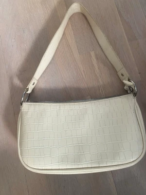 Krämvit axelväska med krokoprägling - Snygg krämvit handväska i avlång modell med croco-mönstrad yta. Väskan har ett kort axelband och stängs med dragkedja upptill. Perfekt för dig som gillar stilrena accessoarer med trendig känsla.